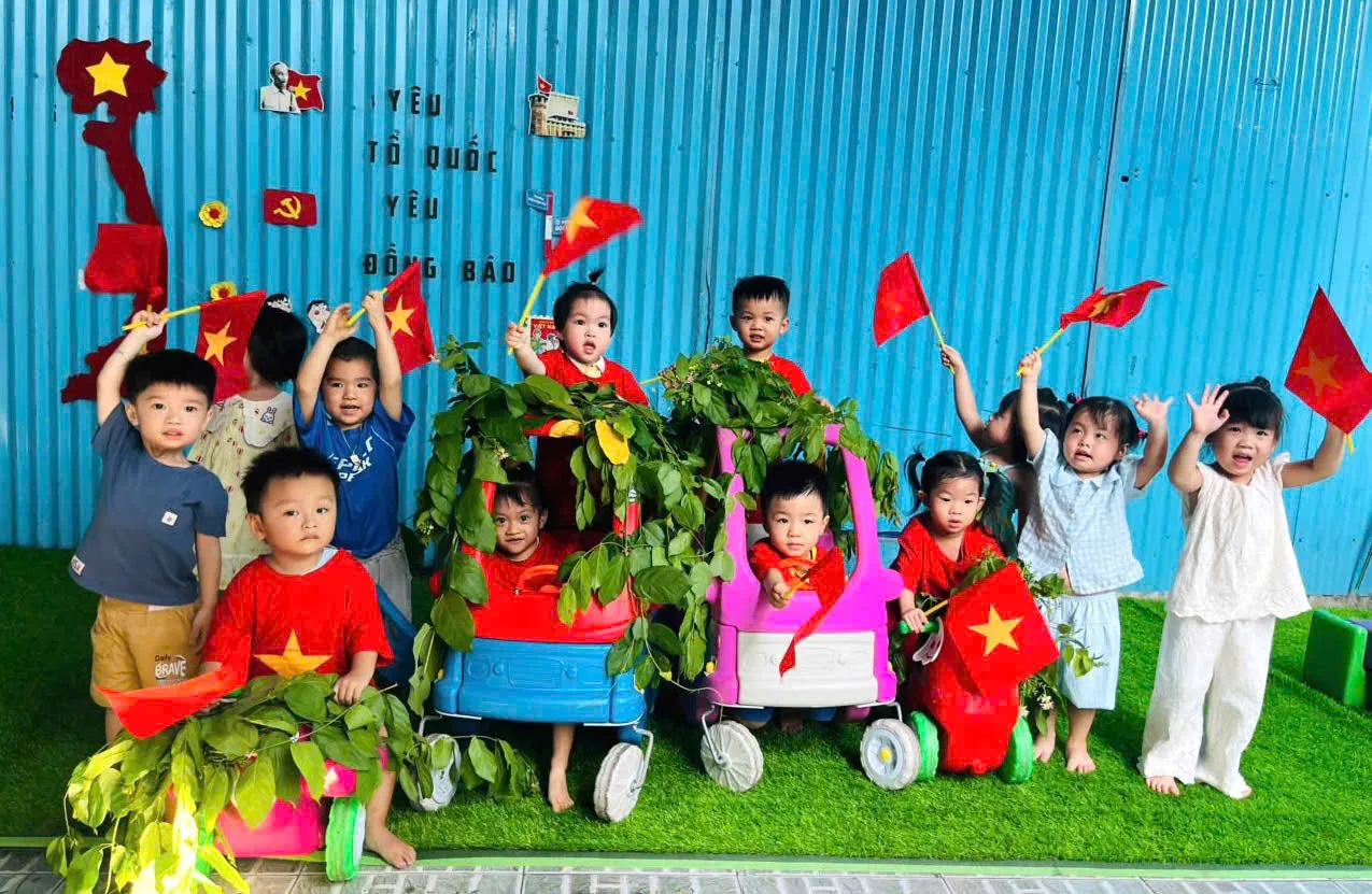 Các bé cầm cờ Tổ quốc vui chơi bên mô hình xe ô tô bìa các-tông